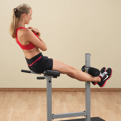 A woman in a red sports bra and black shorts is performing an exercise on a gym machine, focusing on her core while sitting with her legs extended.