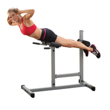 A woman in a red sports bra and black shorts performs an exercise on a gym equipment designed for back extensions, with her legs extended and arms positioned behind her head.