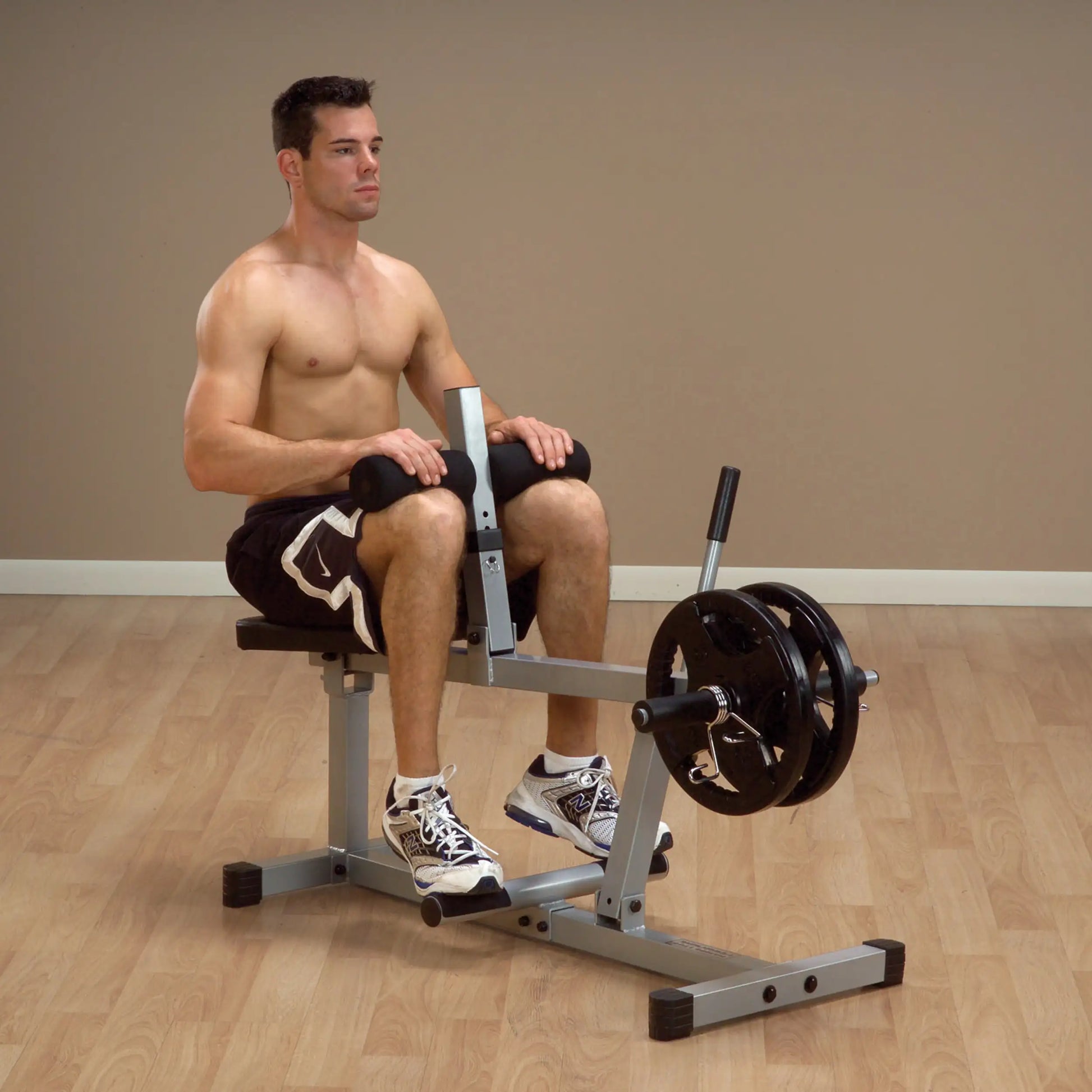 A shirtless man sitting on a leg extension machine in a gym setting, preparing to perform an exercise. He is wearing black shorts and athletic shoes, with weights attached to the machine. The background features a neutral-colored wall and wooden flooring.