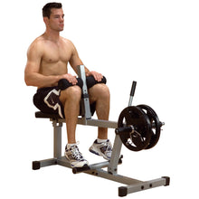 A shirtless man sitting on a leg extension machine in a gym, preparing to perform an exercise with weights attached.