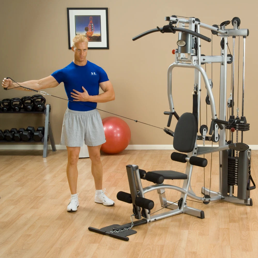 A man in a blue shirt and gray shorts is exercising with a cable machine in a gym. He is performing a stretching motion while holding the cable with one hand. In the background, there are dumbbells and a stability ball.