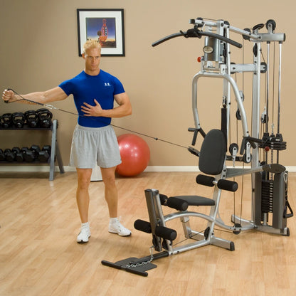 A man in a blue shirt and gray shorts is exercising with a cable machine in a gym. He is performing a stretching motion while holding the cable with one hand. In the background, there are dumbbells and a stability ball.