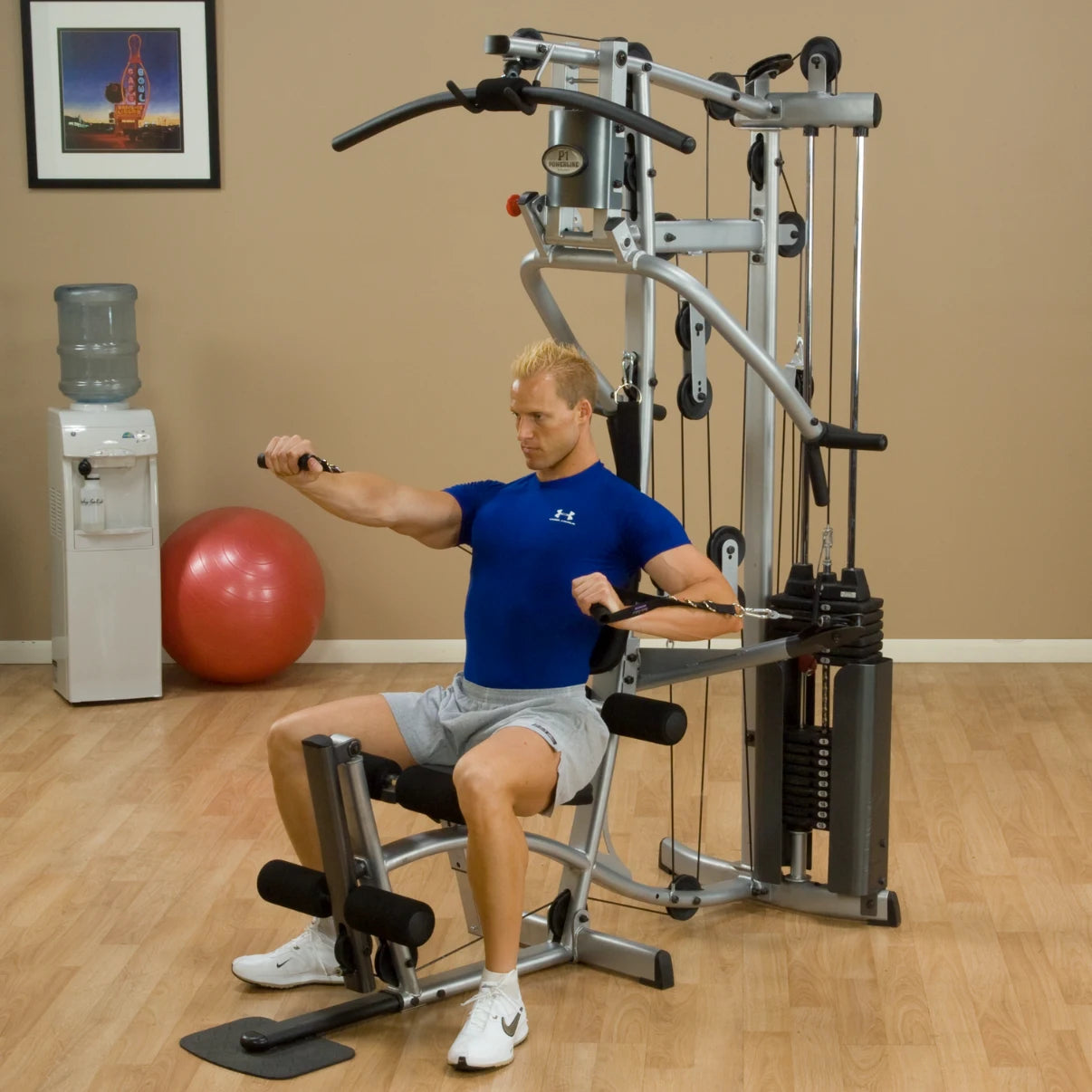 A man in a blue shirt performing a seated exercise on a multi-gym machine in a home gym setting, with a water cooler and exercise ball in the background.