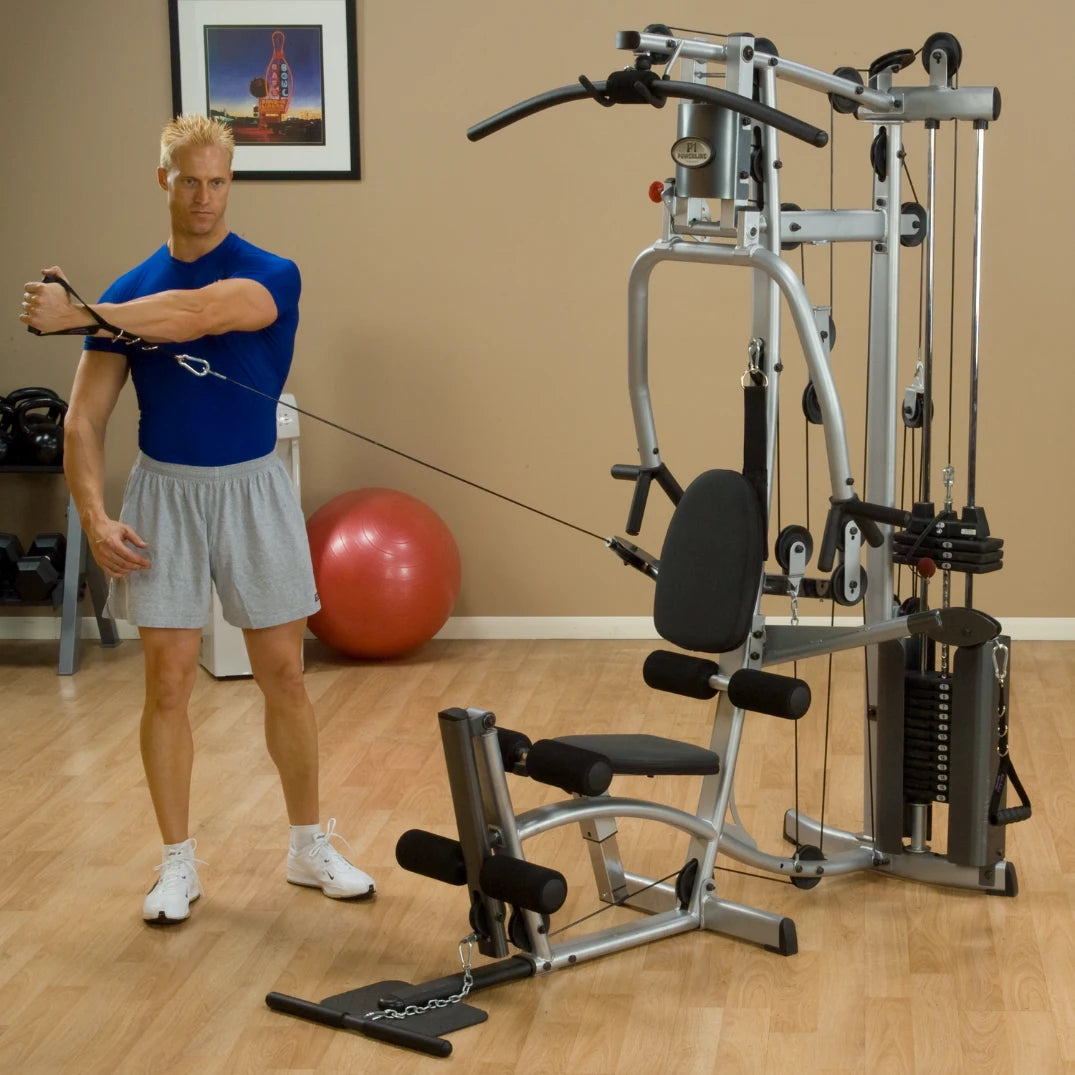 A muscular man wearing a blue shirt and gray shorts is performing a cable exercise on a home gym machine, with a red exercise ball in the background.