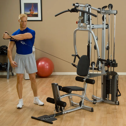A muscular man wearing a blue shirt and gray shorts is performing a cable exercise on a home gym machine, with a red exercise ball in the background.