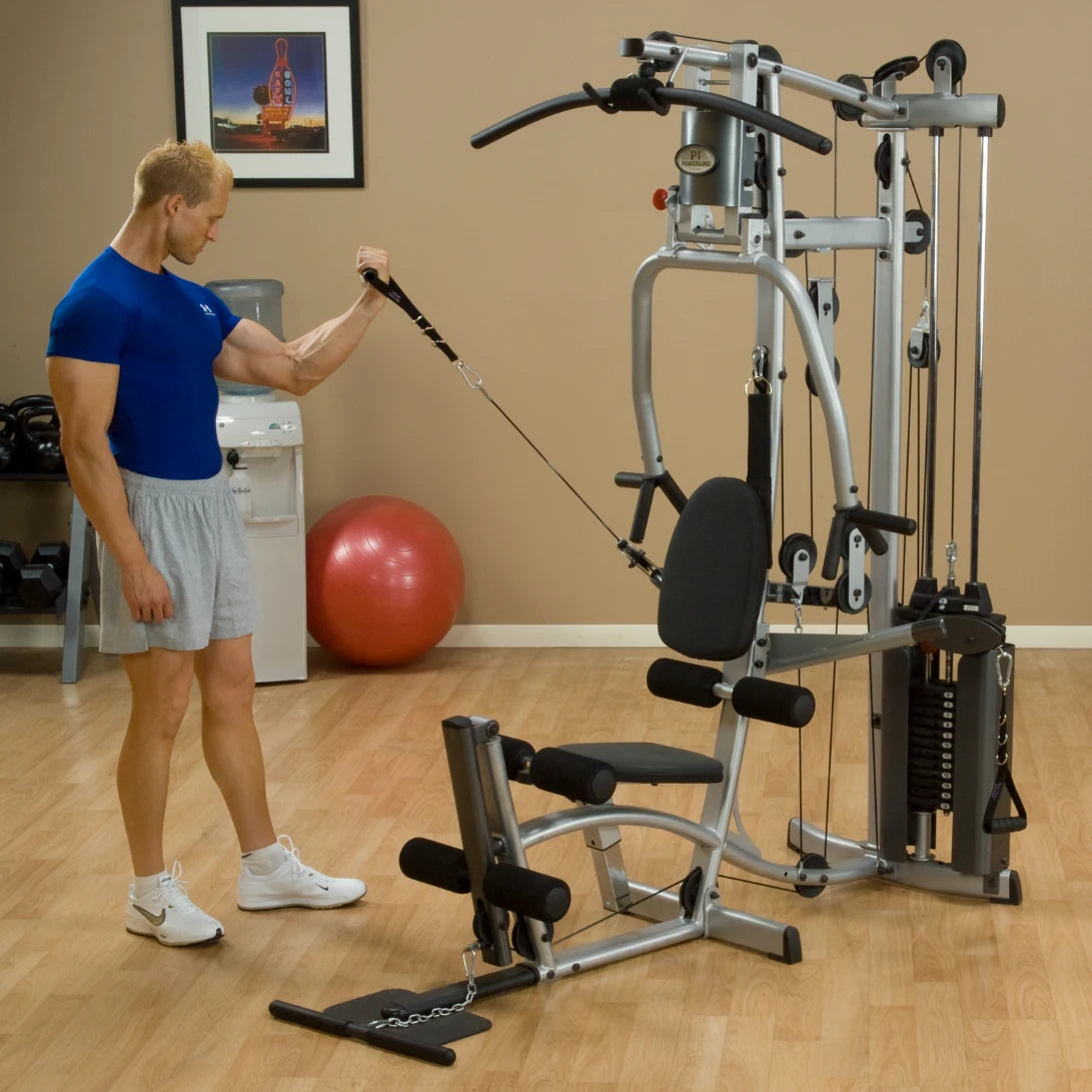 A muscular man in a blue shirt and gray shorts is using a cable attachment on a multi-gym machine in a fitness studio. There is a red exercise ball in the background, along with a water cooler and weights.