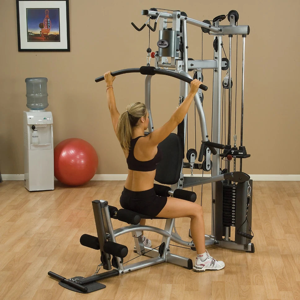 A woman performing a lat pulldown exercise on a home gym machine, wearing a black sports bra and shorts, with a water cooler and exercise ball in the background.