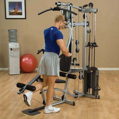 A person in a blue shirt and gray shorts is exercising on a multi-gym machine in a fitness setting, with a water cooler and exercise ball visible in the background.