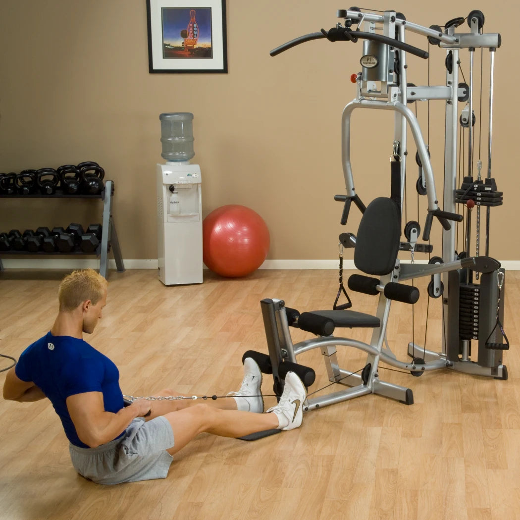 A man exercising on a multi-gym machine in a home gym setting, with a water cooler, exercise ball, and dumbbells in the background.