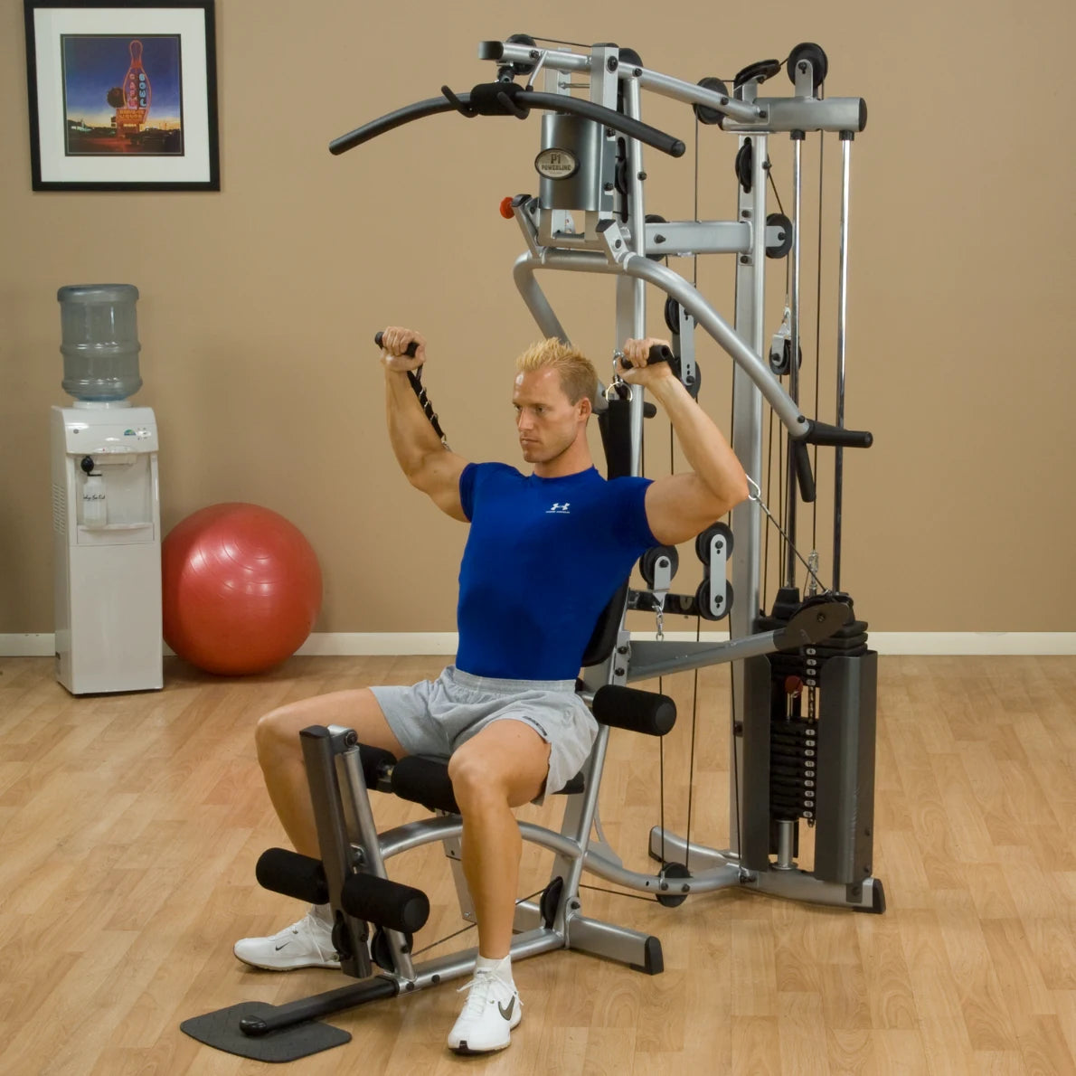 A man in a blue athletic shirt and gray shorts performing a shoulder exercise on a gym machine in a fitness studio, with a water cooler and exercise ball in the background.