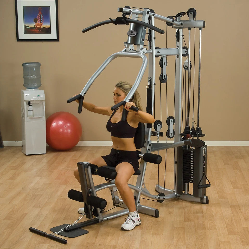 A woman using a multi-gym exercise machine, performing a seated workout in a home gym setting. A water cooler and exercise ball are visible in the background.
