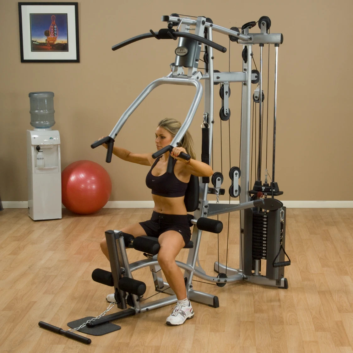 A woman exercising on a multi-gym machine in a home gym setting, with a water cooler and exercise ball in the background.