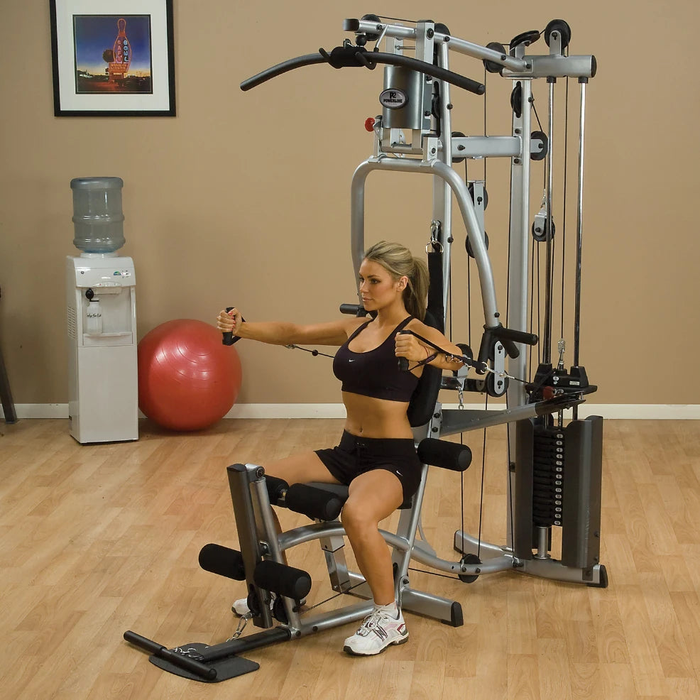 A woman in athletic wear using a multi-gym strength training machine, performing a seated exercise with cables, in a fitness room with a water cooler and an exercise ball in the background.