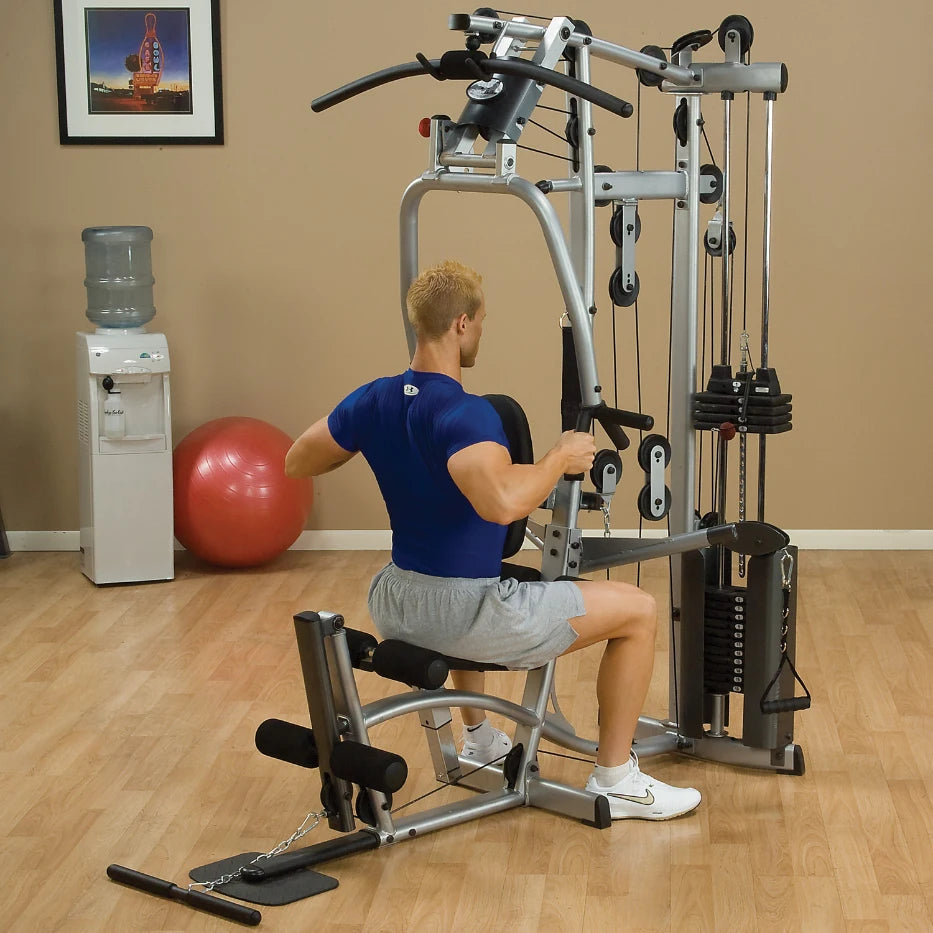 A man exercising on a multi-gym machine in a fitness studio, performing a seated row exercise. A water cooler and exercise ball are visible in the background.