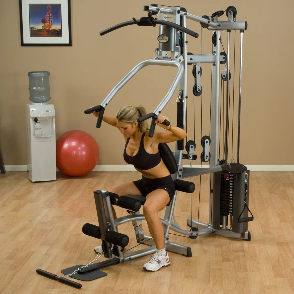A woman exercising on a gym machine in a fitness studio, demonstrating strength training. The background includes a water cooler, a red exercise ball, and a framed picture on the wall.