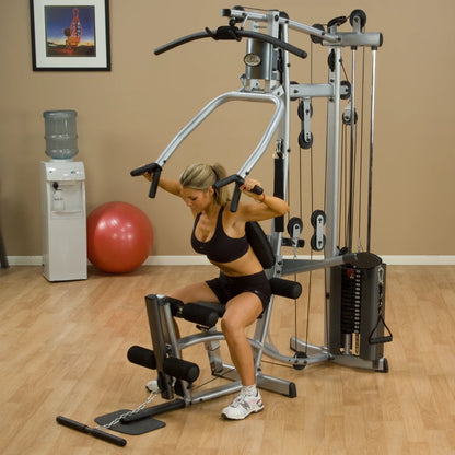 A woman exercising on a gym machine in a fitness studio, demonstrating strength training. The background includes a water cooler, a red exercise ball, and a framed picture on the wall.