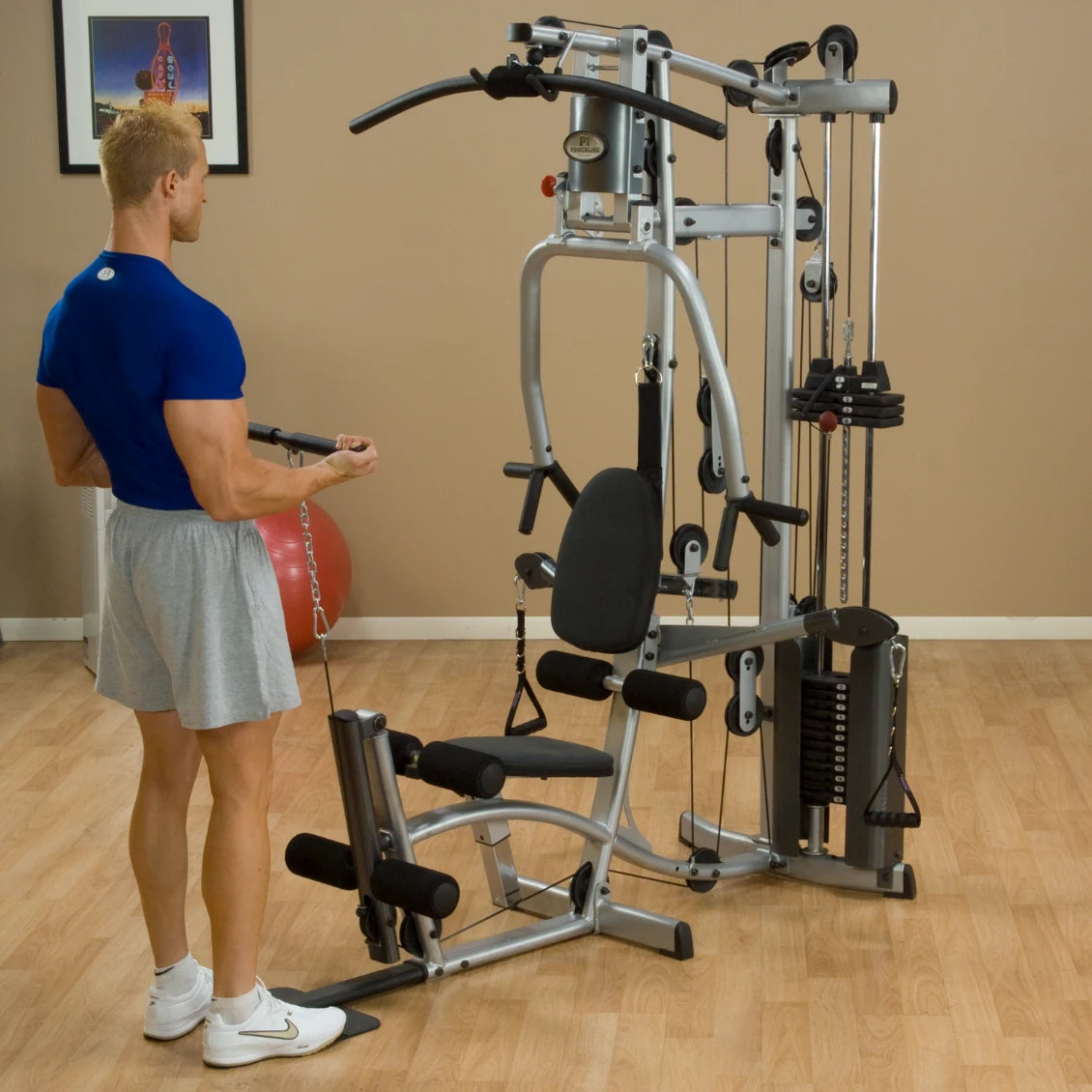 A man in a blue shirt and gray shorts using a multi-gym exercise machine in a home gym setting, with a red exercise ball and framed artwork in the background.