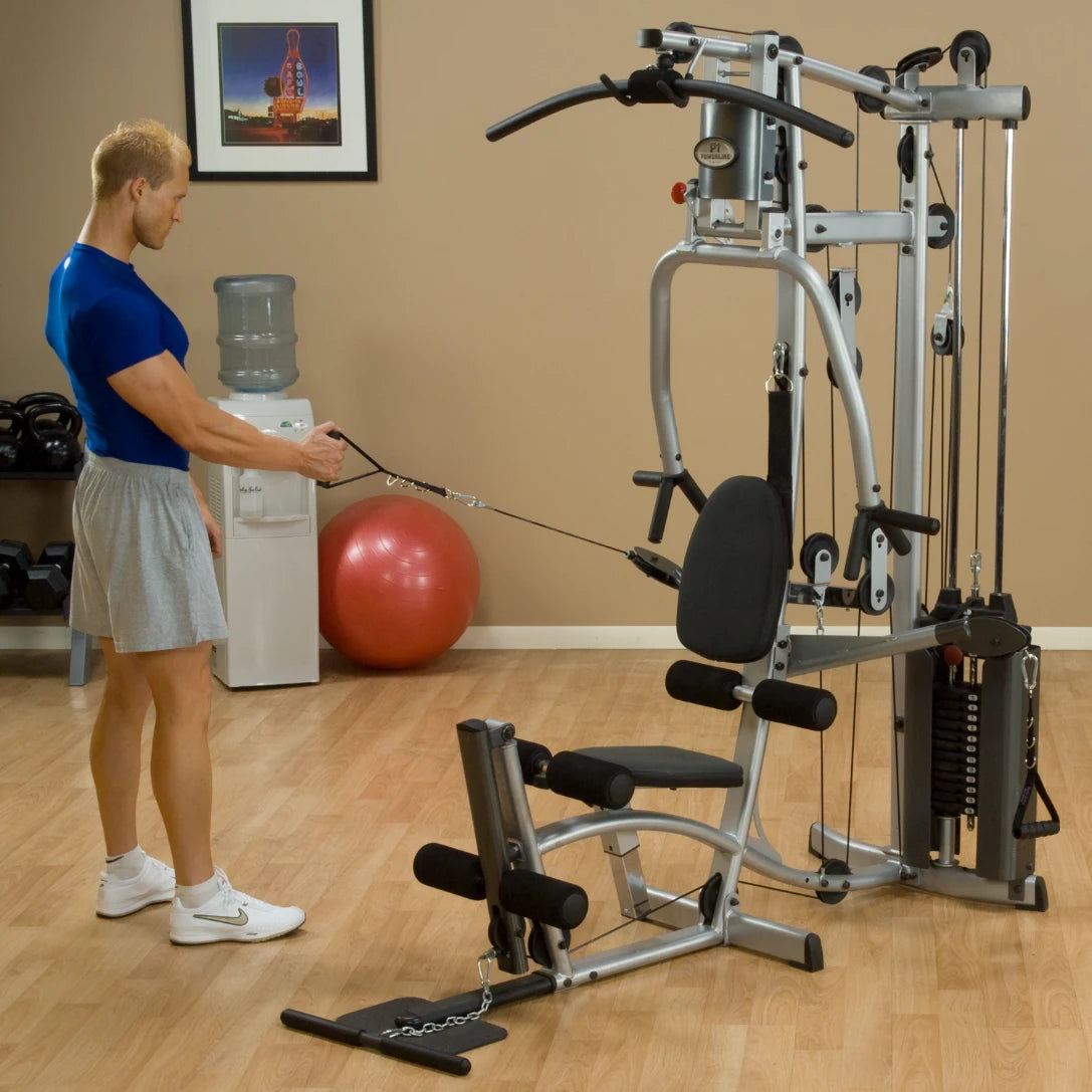 A man in a blue shirt and gray shorts is using a multi-gym machine in a home gym setting. He is pulling a cable attachment while standing beside the machine, which includes a seat and various exercise components. A water cooler and exercise ball are visible in the background.