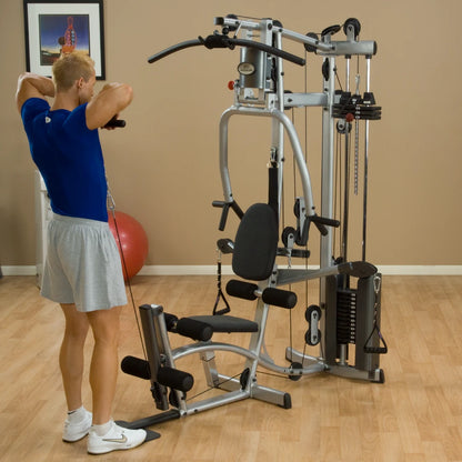 A person exercising on a multi-gym machine, performing a back row exercise, in a well-lit room with fitness equipment and a large exercise ball in the background.