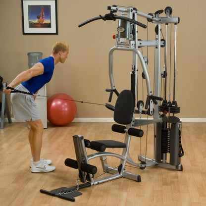 A man in a blue shirt and gray shorts performs a cable row exercise on a multi-gym machine in a fitness studio, with a red exercise ball in the background.