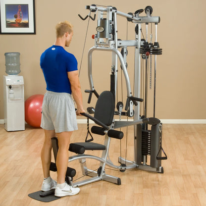 A man wearing a blue shirt and gray shorts is exercising on a multi-gym machine in a bright indoor gym setting, with a water cooler and exercise ball in the background.