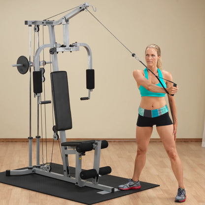 A woman in a turquoise sports bra and black shorts is performing an exercise using a cable machine in a gym setting. The machine features a seat, a vertical bar, and cables for resistance training. The background is a light-colored wall with a wooden floor.