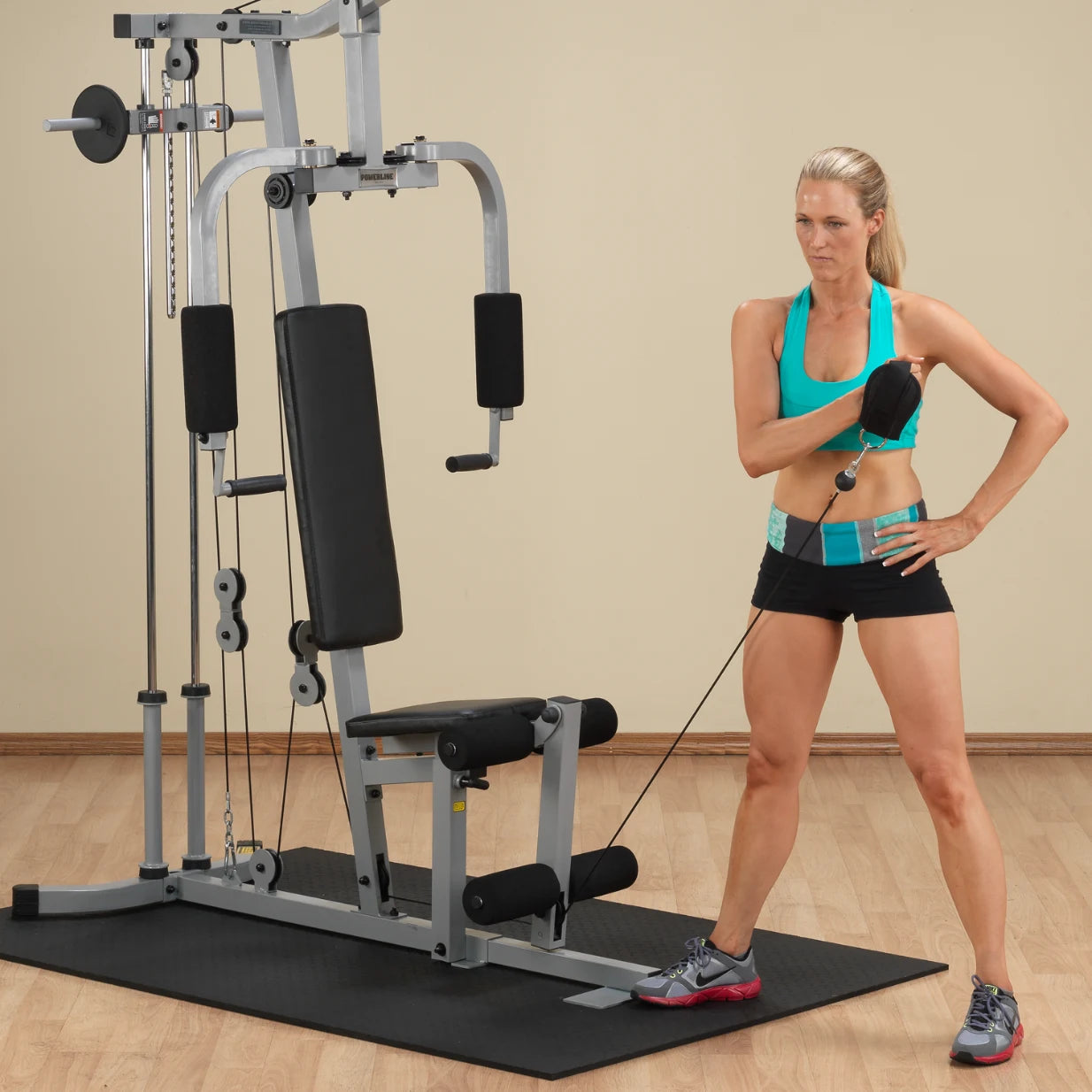 A woman in a turquoise athletic top and black shorts is using a fitness machine, performing a cable exercise. She appears focused, with one hand on her hip and the other holding a cable attachment. The gym has a wooden floor and a neutral background.