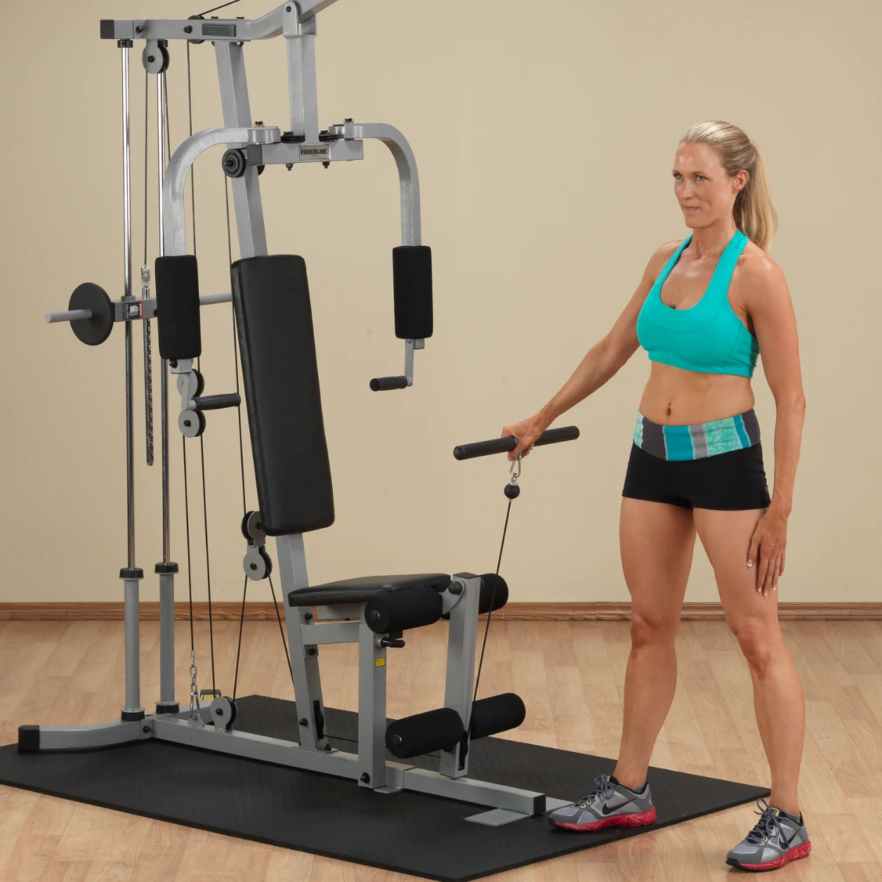 A woman in a turquoise sports bra and black shorts stands beside a multi-gym exercise machine, holding a cable handle with one hand, preparing for a workout in a well-lit room.