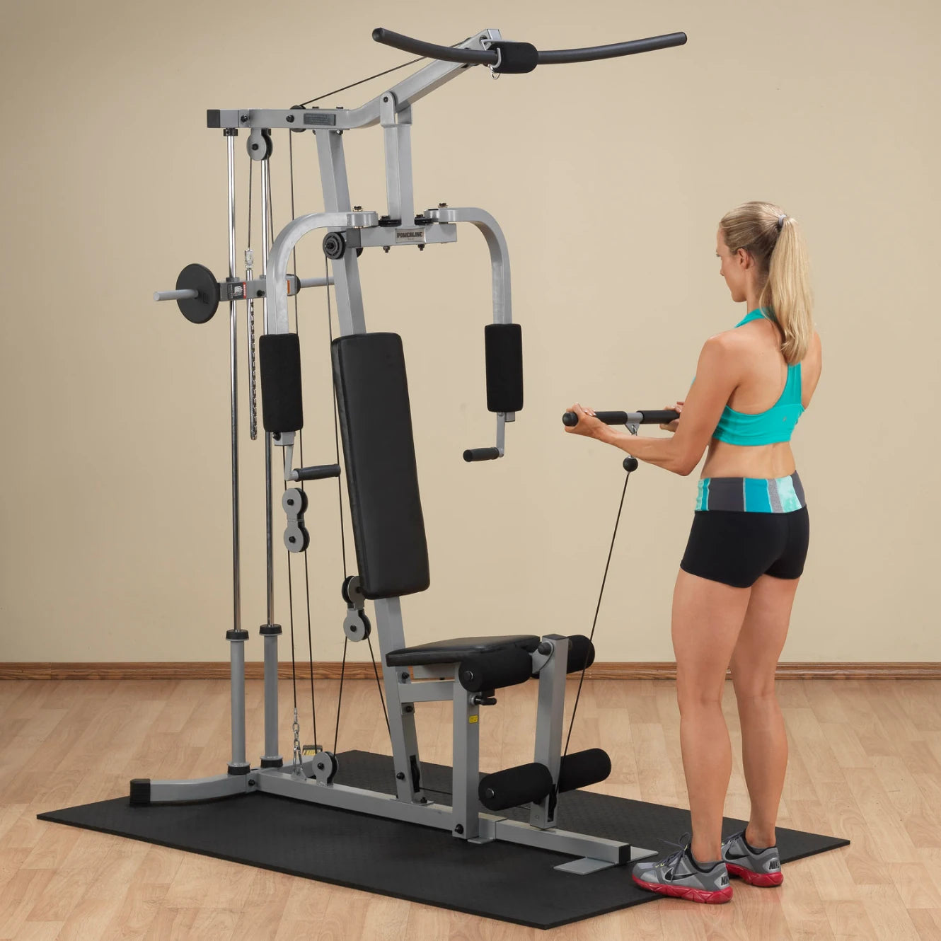 A woman in a turquoise sports bra and black shorts is using a multi-gym exercise machine, performing a cable exercise. The setting features a light-colored wall and wooden flooring.