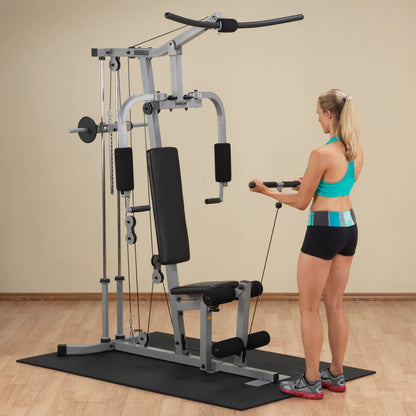 A woman in a turquoise sports bra and black shorts is using a multi-gym exercise machine, performing a cable exercise. The setting features a light-colored wall and wooden flooring.
