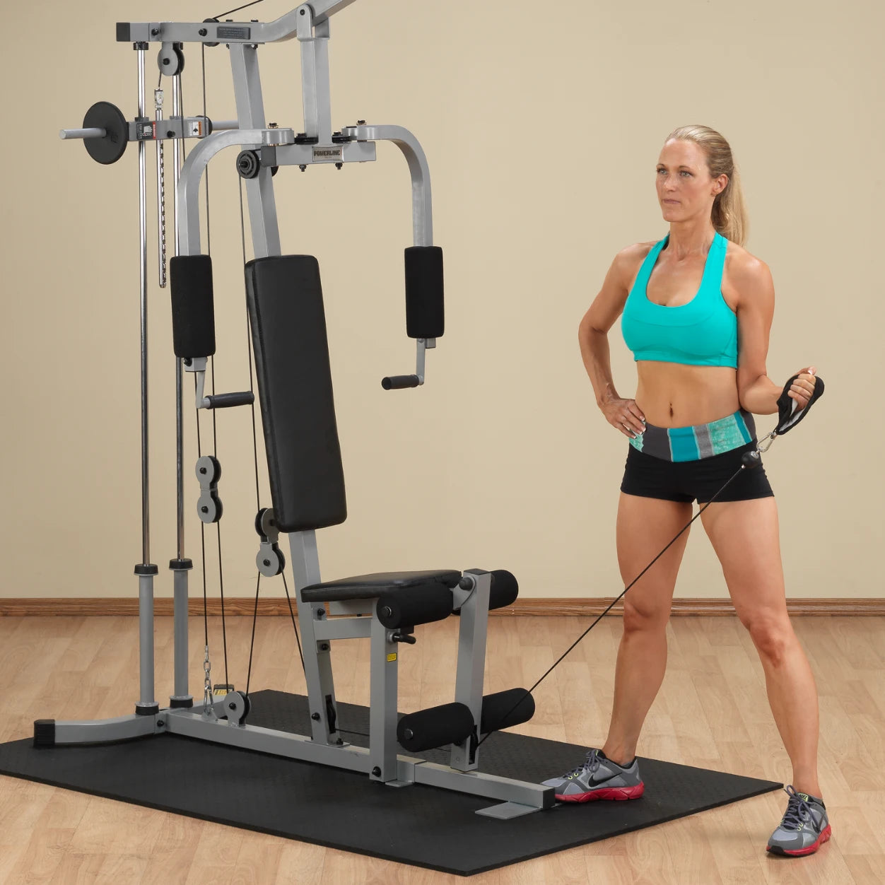A woman in a turquoise sports bra and black shorts stands next to a multi-gym exercise machine, holding a resistance cable with one hand while resting her other hand on her hip.