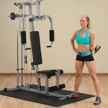 A woman in a turquoise sports bra and black shorts stands next to a multi-gym exercise machine, holding a resistance cable with one hand while resting her other hand on her hip.