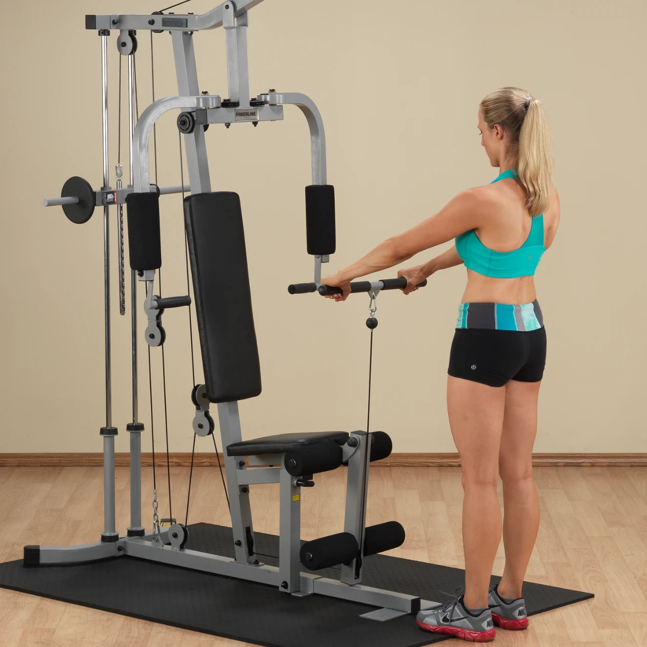 A woman in a turquoise sports bra and black shorts using a cable exercise machine in a gym setting.