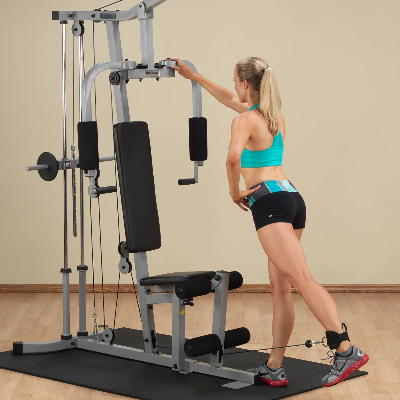 A woman in athletic wear using a multi-gym machine, performing an exercise with a cable attachment while standing on a gym mat.