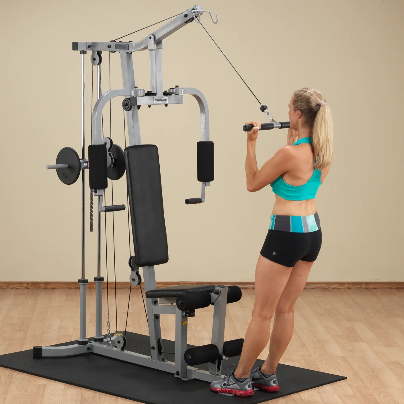 A woman in a turquoise sports bra and black shorts using a cable machine for strength training in a gym setting.