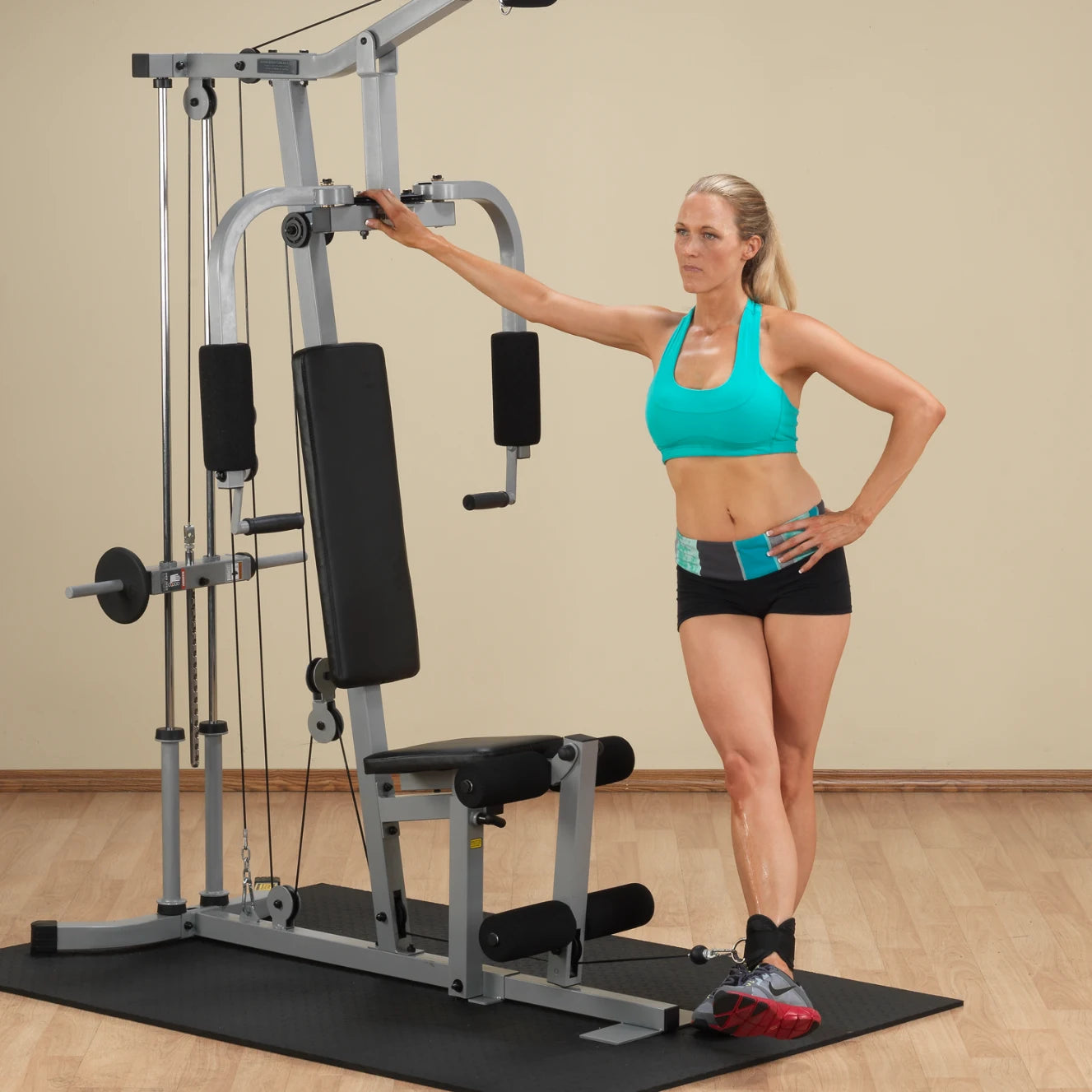 A fit woman in a turquoise sports bra and black shorts stands next to a multi-gym workout machine, leaning on it with one arm while posing confidently. The background features a neutral wall and wooden flooring.