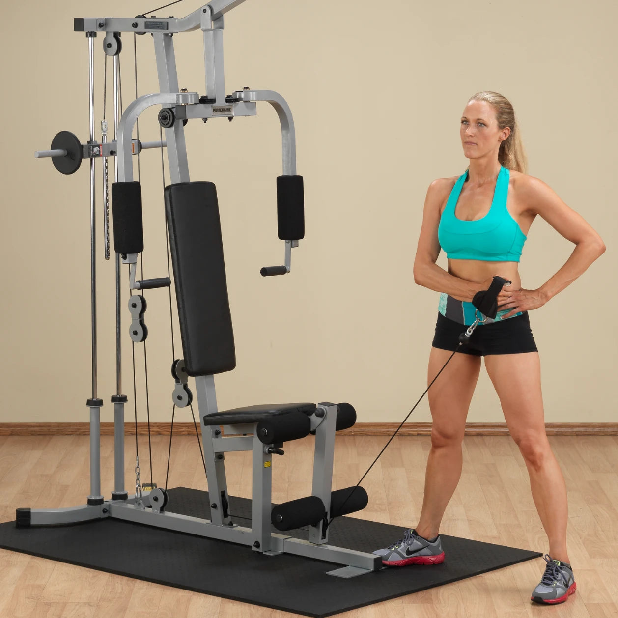 A fit woman in a turquoise sports bra and black shorts stands beside a multi-gym workout machine, holding a resistance cable with one hand on her hip, preparing to exercise.