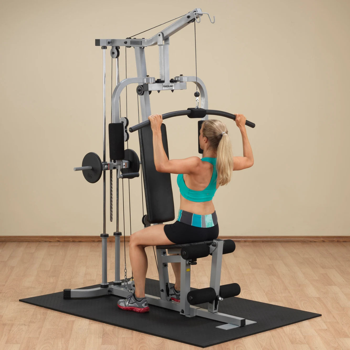 A woman with long blonde hair wearing a teal sports bra and black shorts exercises on a weight machine, performing a lat pull-down in a gym setting with a light-colored wall and wooden floor.