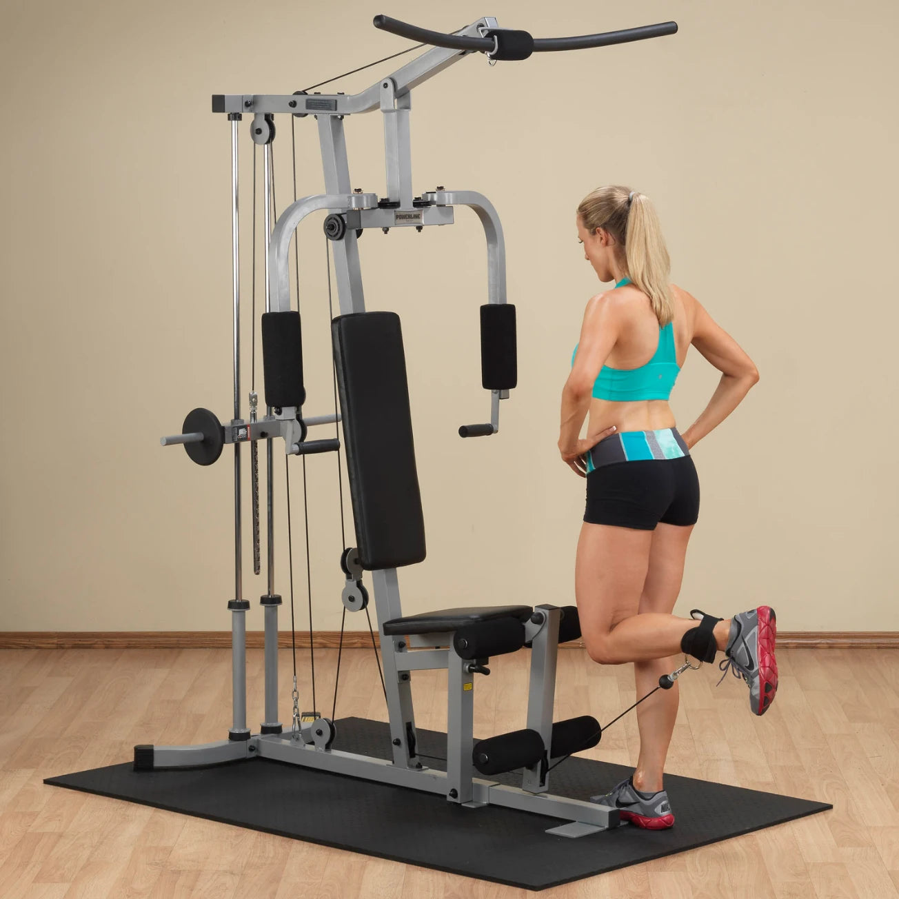 A woman in a turquoise sports bra and black shorts is using a multi-gym machine in a home workout setting, focusing on her leg exercise while standing on a black mat.