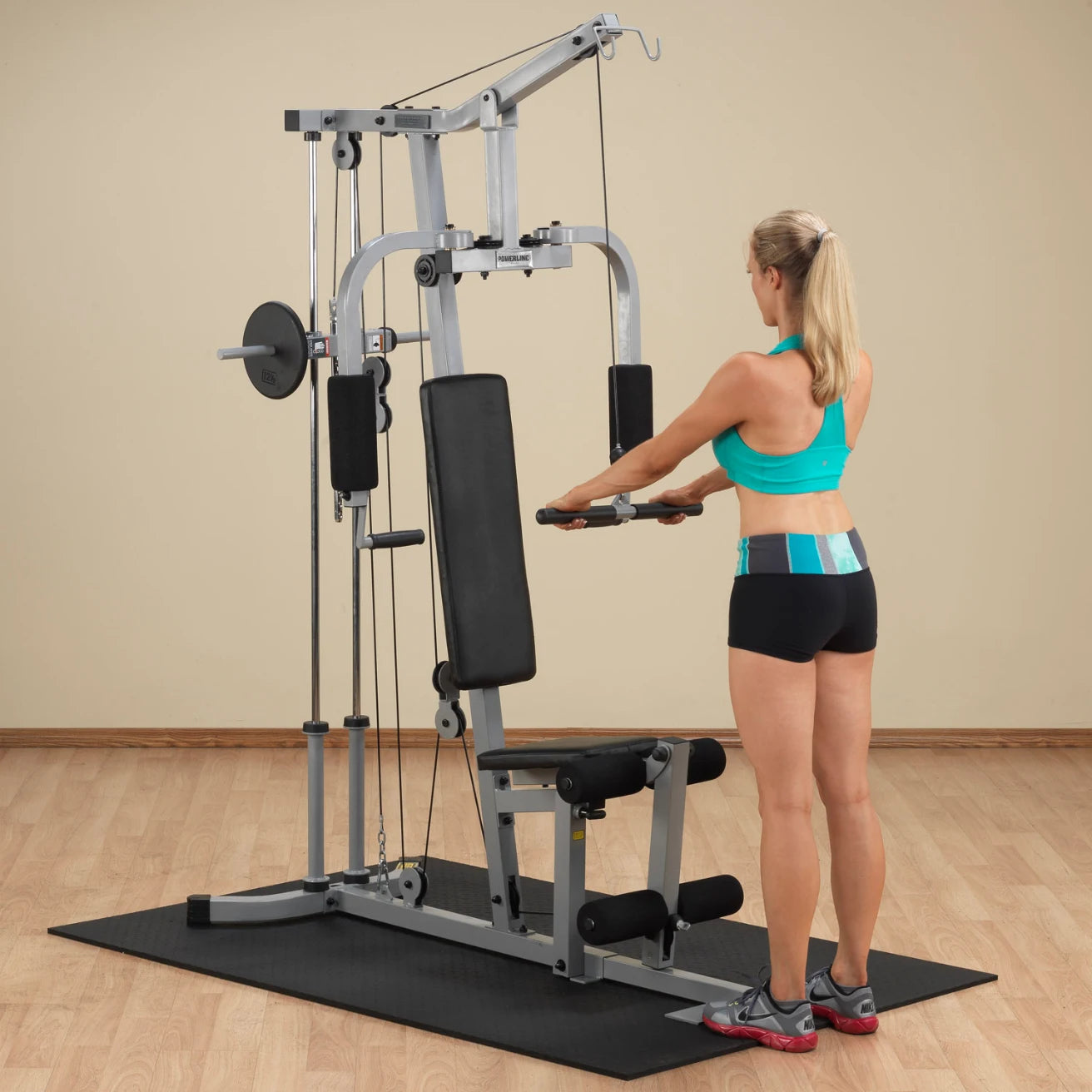A woman in a turquoise sports bra and black shorts is using a multi-gym workout machine, focusing on her upper body exercise. The gym is equipped with a rubber mat and wooden flooring, providing a fitness environment.