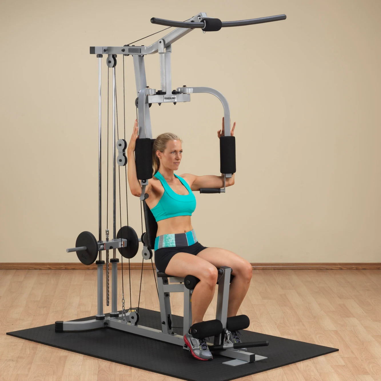 A woman in a turquoise sports bra and black shorts is seated on a multi-gym machine, performing an exercise with her arms raised, demonstrating strength training in a well-lit gym environment.