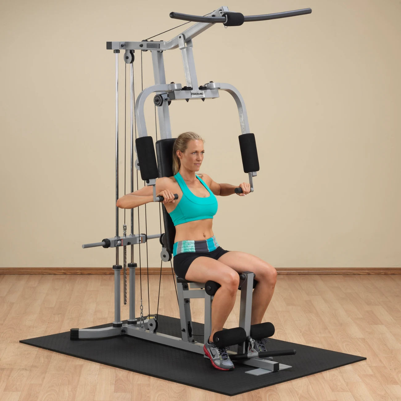 A woman performing an exercise on a multi-gym machine, wearing a turquoise sports bra and black shorts, with her hands gripping the machine's handles in a seated position. The gym is equipped with a black mat and wooden flooring.