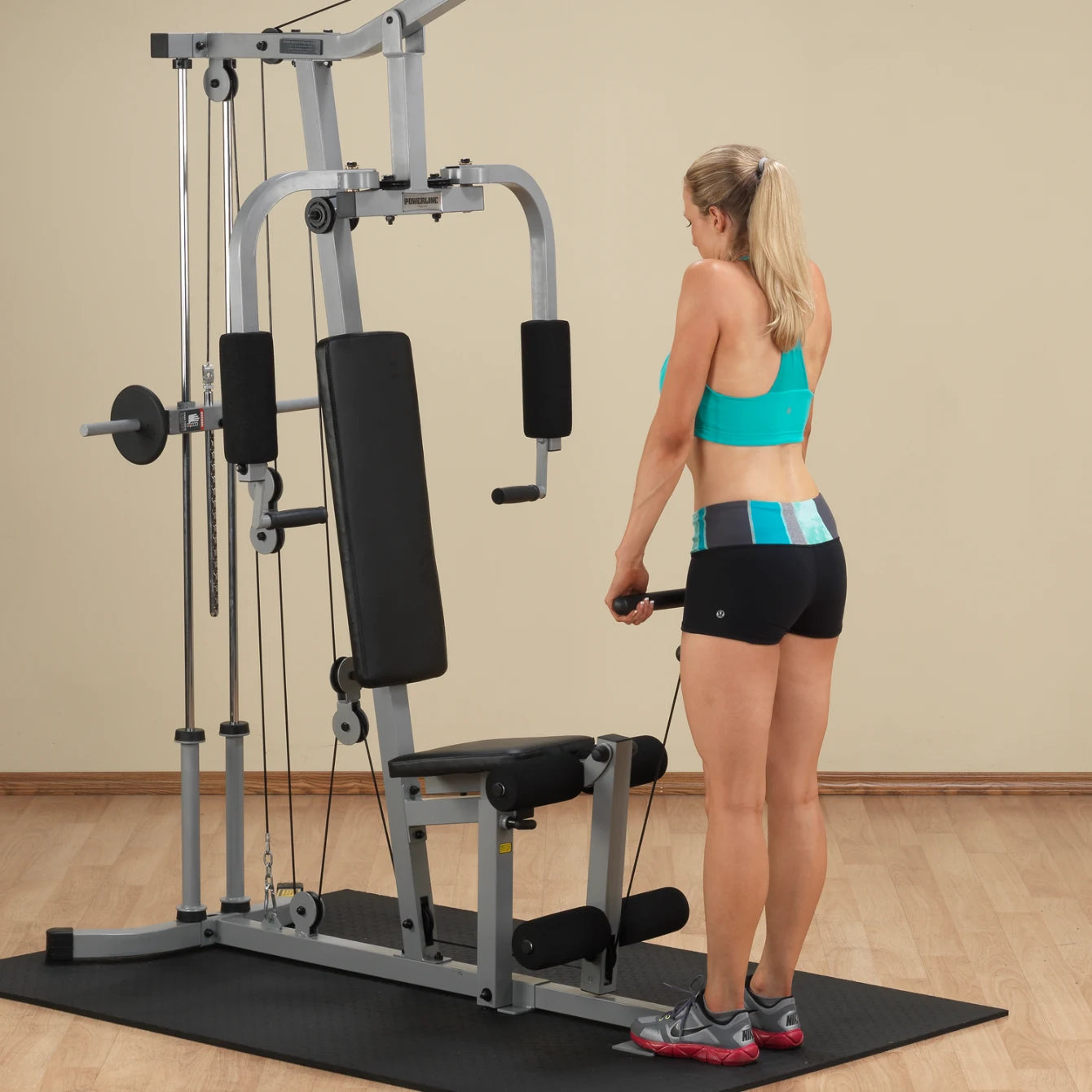 A woman in a turquoise sports bra and black shorts is using a cable machine for exercise in a gym setting. The machine features a bench and various pulleys, with a rubber mat on the floor.