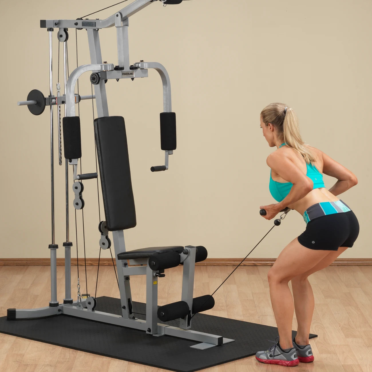 A woman wearing a turquoise sports bra and black shorts is using a cable machine for strength training in a gym setting. The machine features a seat and various pulleys.