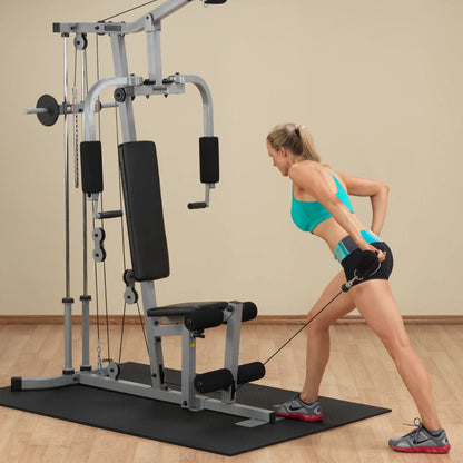 A woman in a turquoise sports bra and black shorts performs a cable row exercise using a weight machine in a home gym setting.