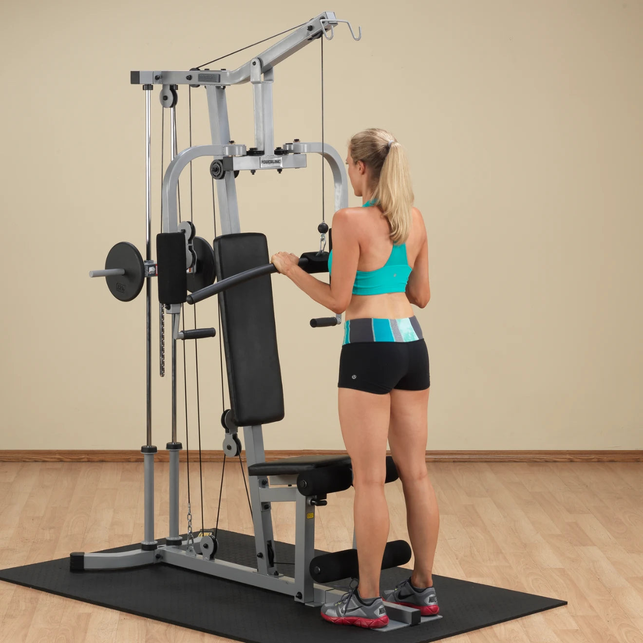 A woman in a turquoise sports bra and black shorts is using a multi-gym machine for strength training in a home gym setting. The background features light-colored walls and wooden flooring.