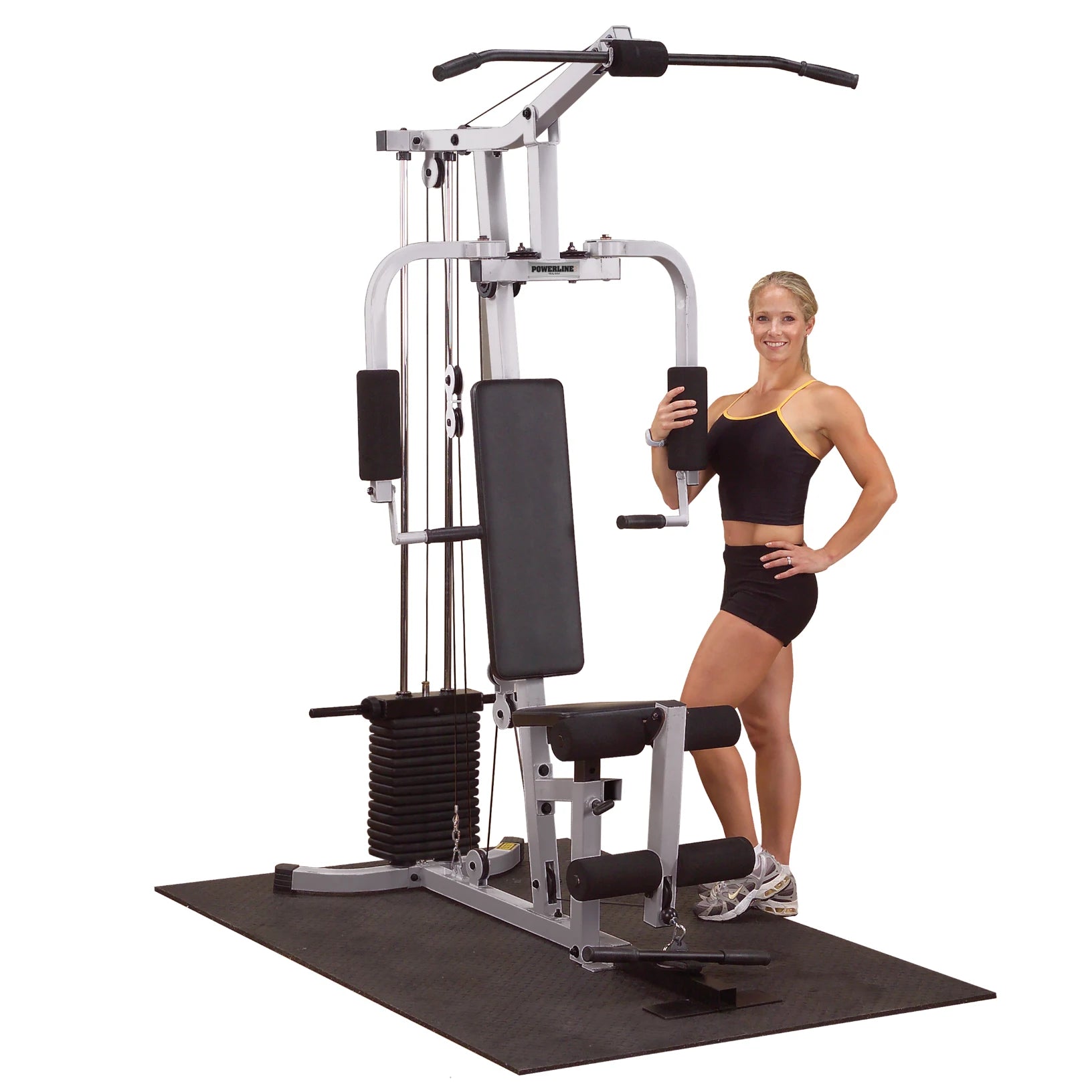 A fit woman in athletic wear stands next to a multi-gym exercise machine, smiling and holding a weight attachment, with a rubber mat underneath.