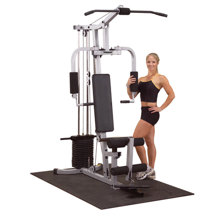 A fit woman in athletic wear stands next to a multi-gym exercise machine, smiling and holding a weight attachment, with a rubber mat underneath.