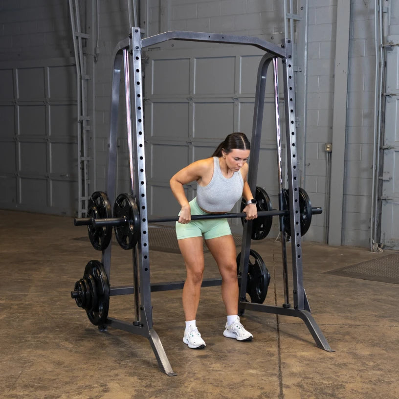 A woman performing a bent-over row exercise with a barbell in a gym setting, using a squat rack with weights on either side. She is wearing a gray tank top and green shorts, focused on her form.
