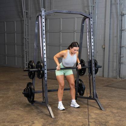 A woman performing a bent-over row exercise with a barbell in a gym setting, using a squat rack with weights on either side. She is wearing a gray tank top and green shorts, focused on her form.